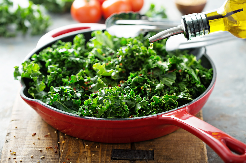 Foods That Can Help Relieve Your Chronic Pain; Quickly sauteed kale with chili flakes in a cast iron pan with olive oil pouring over, healthy cooking concept