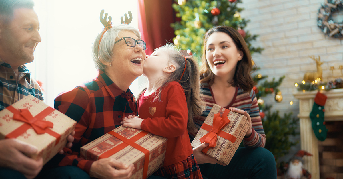 Merry Christmas and Happy Holidays! Grandma, grandpa, mum and child exchanging gifts. Parents and daughters having fun near tree indoors. Loving family with presents in room; blog: Living with Alzheimer’s: How to Enjoy the Holidays