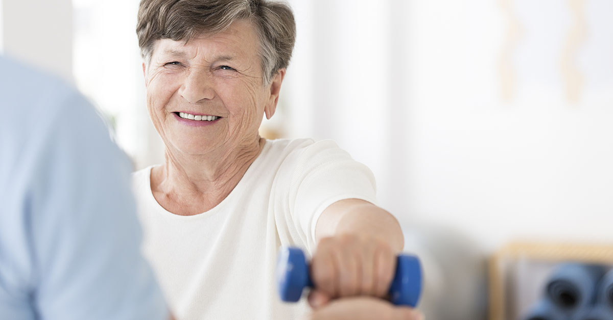 Happy senior woman exercising arms with small weights with help from a physiotherapist; blog: What You Need to Know About Stroke Recovery