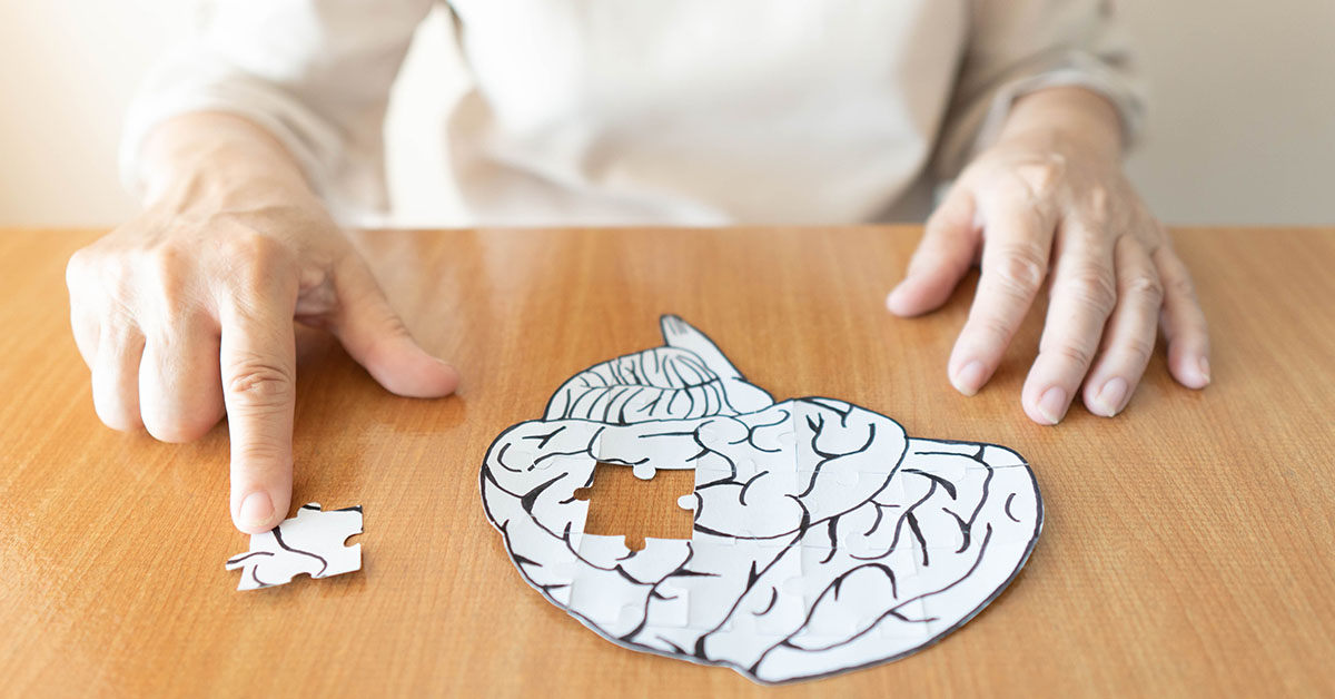 Elderly woman hands putting missing white jigsaw puzzle piece down into the place as a human brain shape. Creative idea for memory loss, dementia, Alzheimer's disease and mental health concept; blog: 10 Early Signs of Alzheimer’s Disease